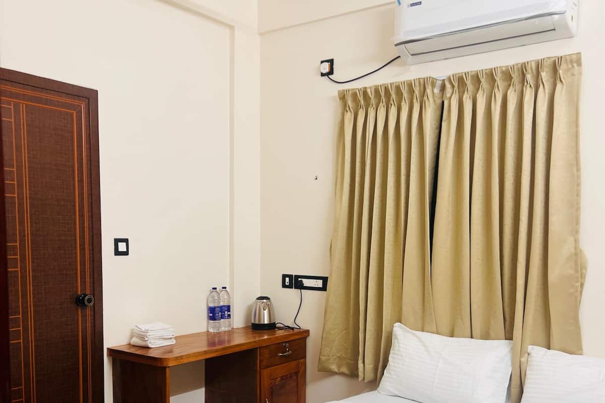 A hotel room with a bed, a wooden wardrobe, a desk with a kettle, a water bottle, and an air conditioning unit.