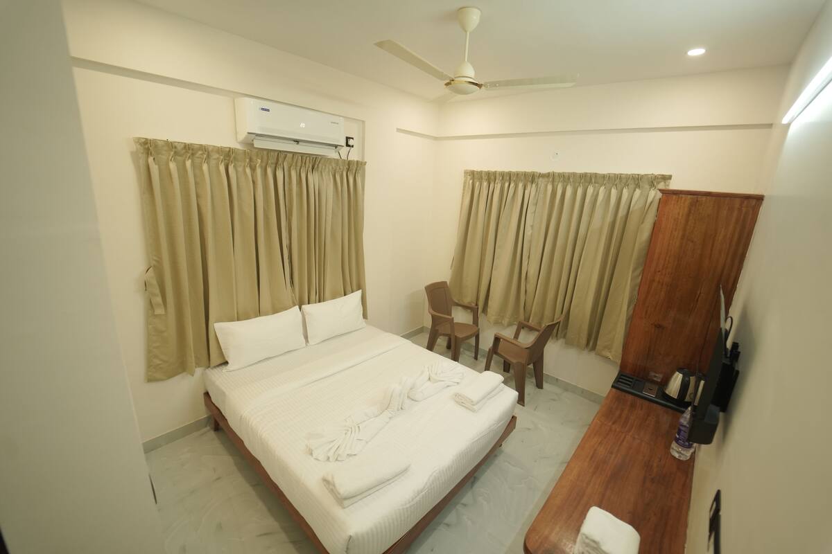 A hotel room with a bed, two chairs, a desk, an air conditioning unit, a ceiling fan, and a wooden wardrobe.