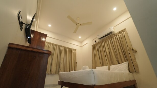 A bedroom with a bed, wooden dresser, ceiling fan, air conditioning unit, and curtains. - Vallimony Luxury Stays-Padmanabha Temple (Thiruvananthapuram)