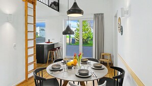 Zona pranzo moderna con un tavolo rotondo, sedie nere e una lampada a sospensione centrale. Si notano in secondo piano una scala in legno e un angolo cottura.