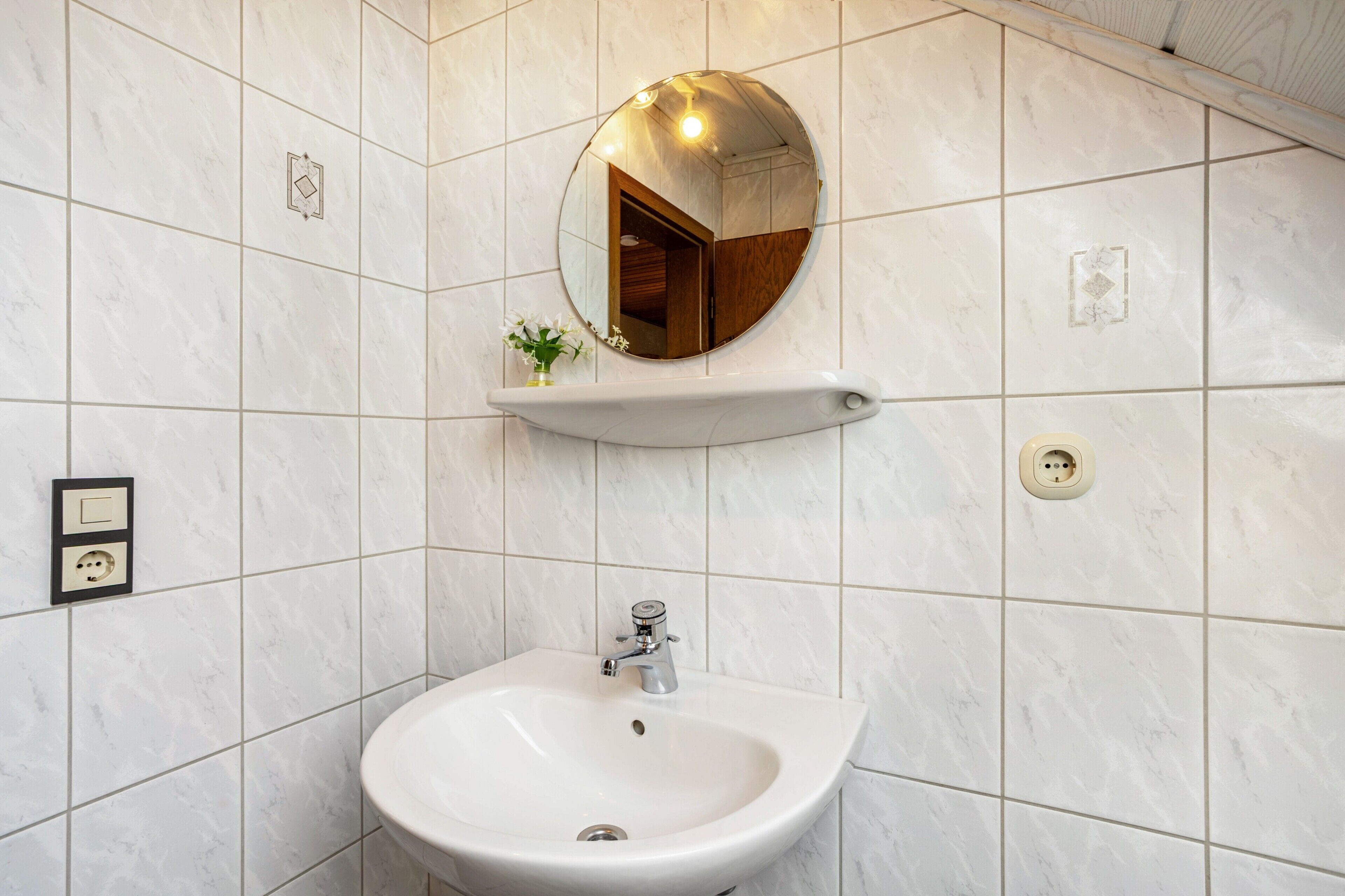 Ein Badezimmer mit einem weißen Waschbecken, einem Spiegel und einem kleinen Ablagefach mit einer Vase.