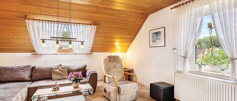 Ein gemütliches Wohnzimmer mit Holzboden, einem Glastisch, einem braunen Sofa, einem beigen Sessel und einem Fenster mit weißen Vorhängen.