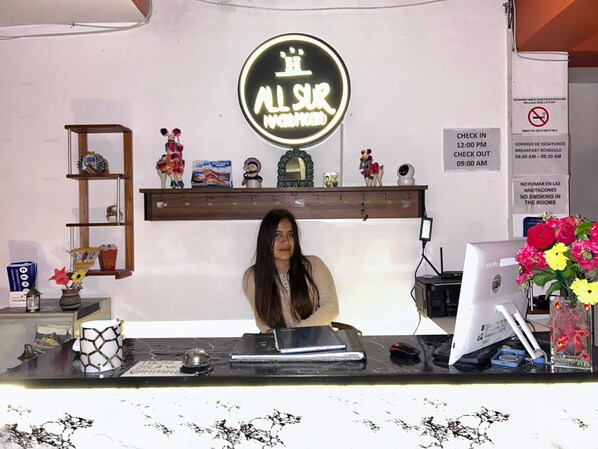Bancada de recepção de hotel com um letreiro de neon que diz "AL SUR", um computador, um telefone e vários objetos decorativos em prateleiras.