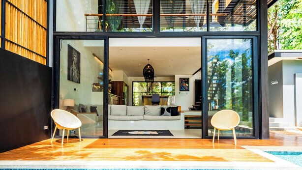 Una casa moderna con puerta de vidrio que da a una sala de estar, piscina y terraza de madera.
