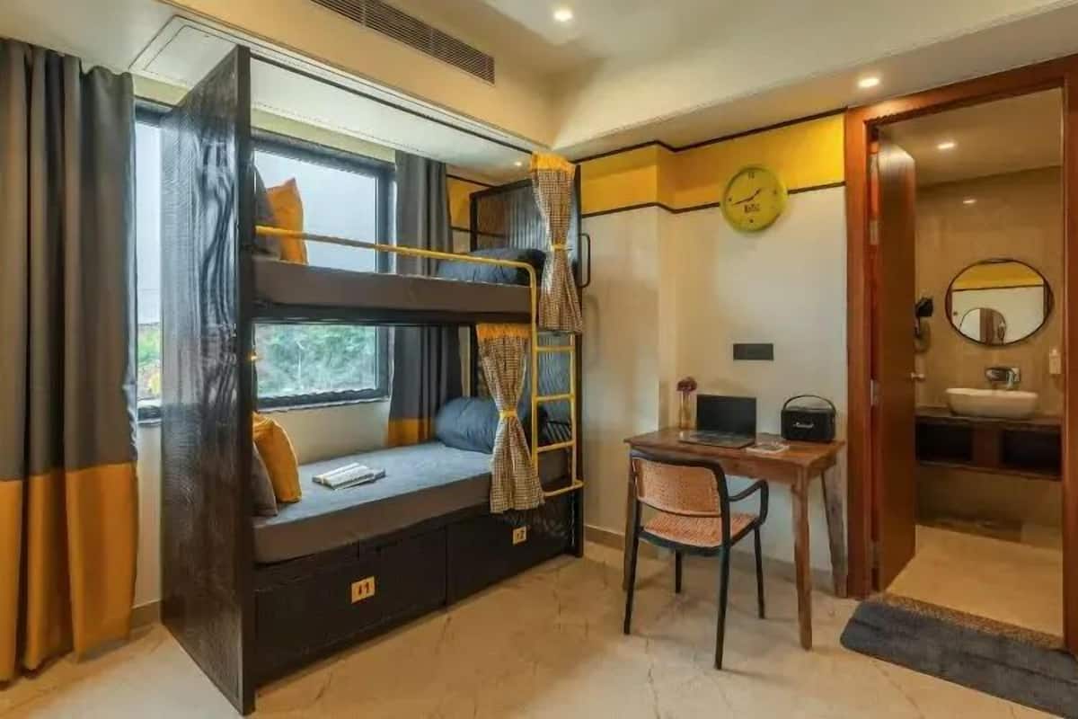 Habitación pequeña con literas, un escritorio, una silla, una mesita, un espejo y baño con lavamanos e inodoro.