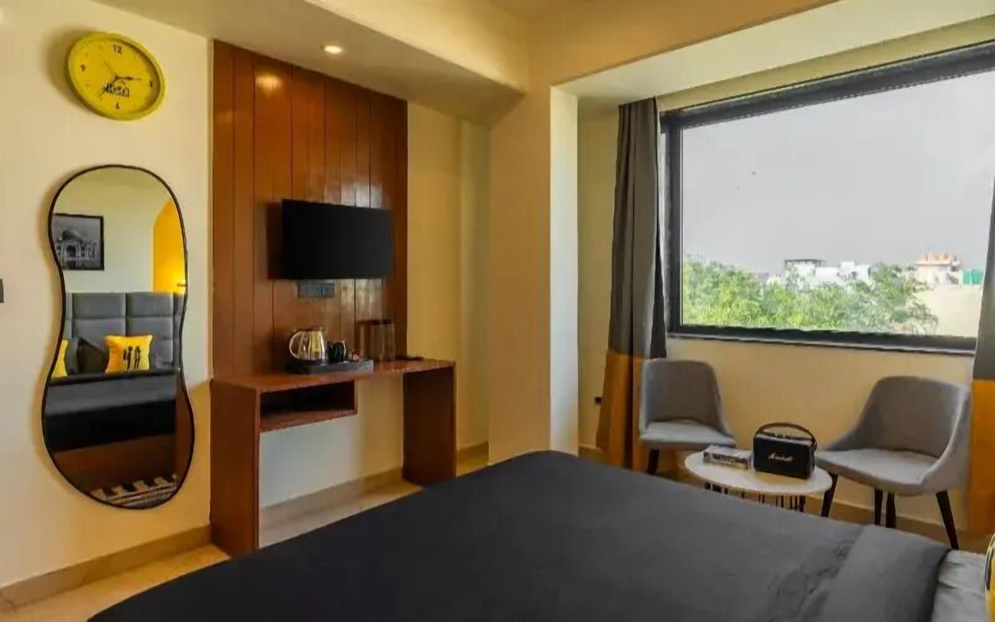 A hotel room with a bed, a TV mounted on the wall, a desk, two chairs, a large window, and a mirror.