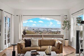 A living room with a large arched window offering a city view, a sofa with decorative pillows, and a small table with books and a vase. - Spanish Manor by Coastline Vacation Rentals (San Diego)