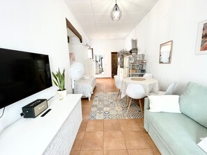 Ein Wohnzimmer mit Fernseher, Ventilator, einer Couch, einem Esstisch mit Stühlen und einer Küche im Hintergrund.