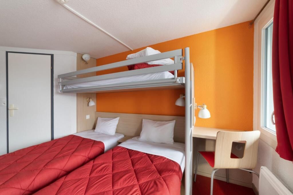 Une chambre d’hôtel compacte avec un lit superposé, un bureau avec une chaise et une fenêtre avec des rideaux rouges.