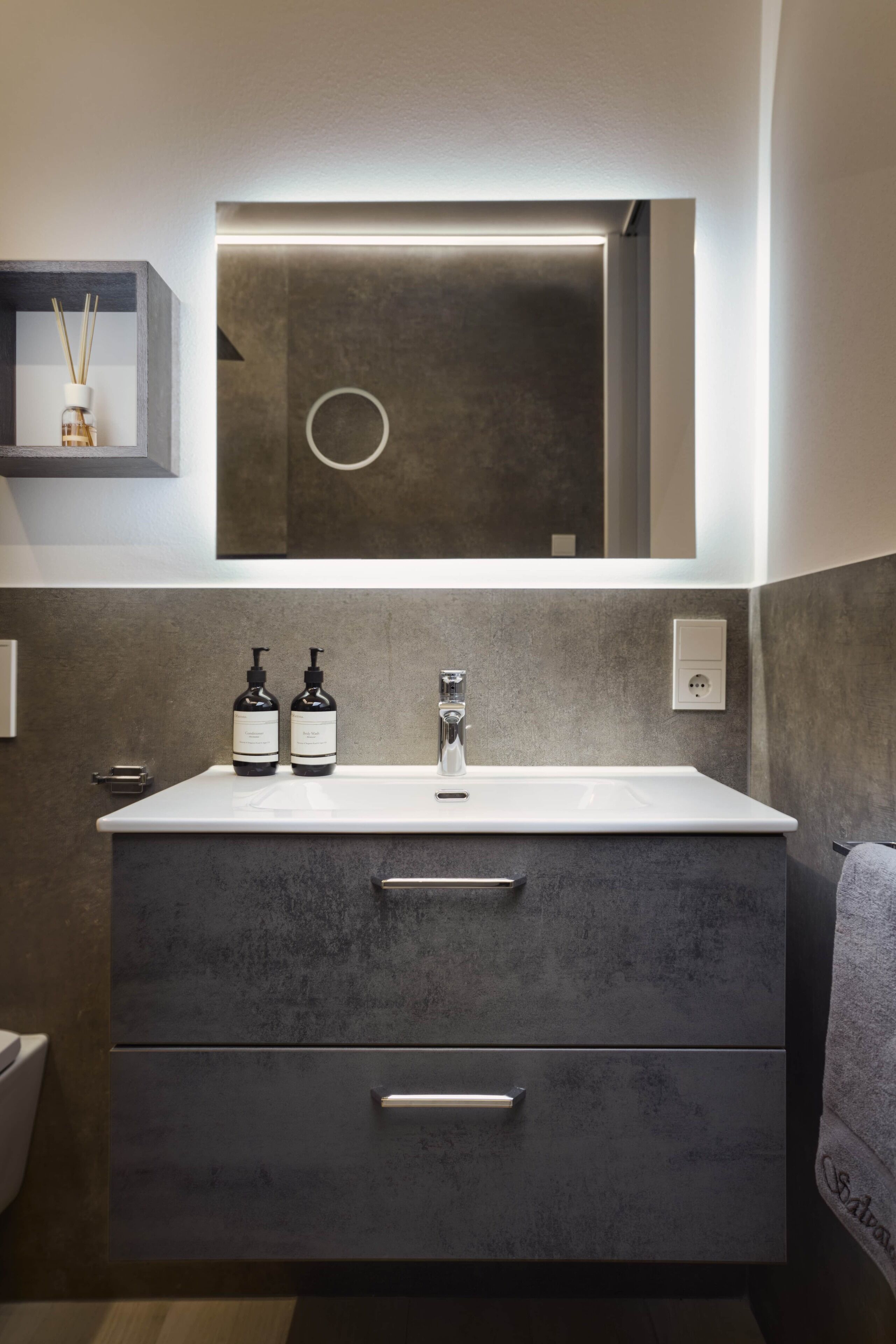 Une salle de bain moderne avec un lavabo blanc, un meuble de couleur gris foncé et un miroir au-dessus. On peut voir deux bouteilles sur le lavabo et un porte-serviettes à droite.