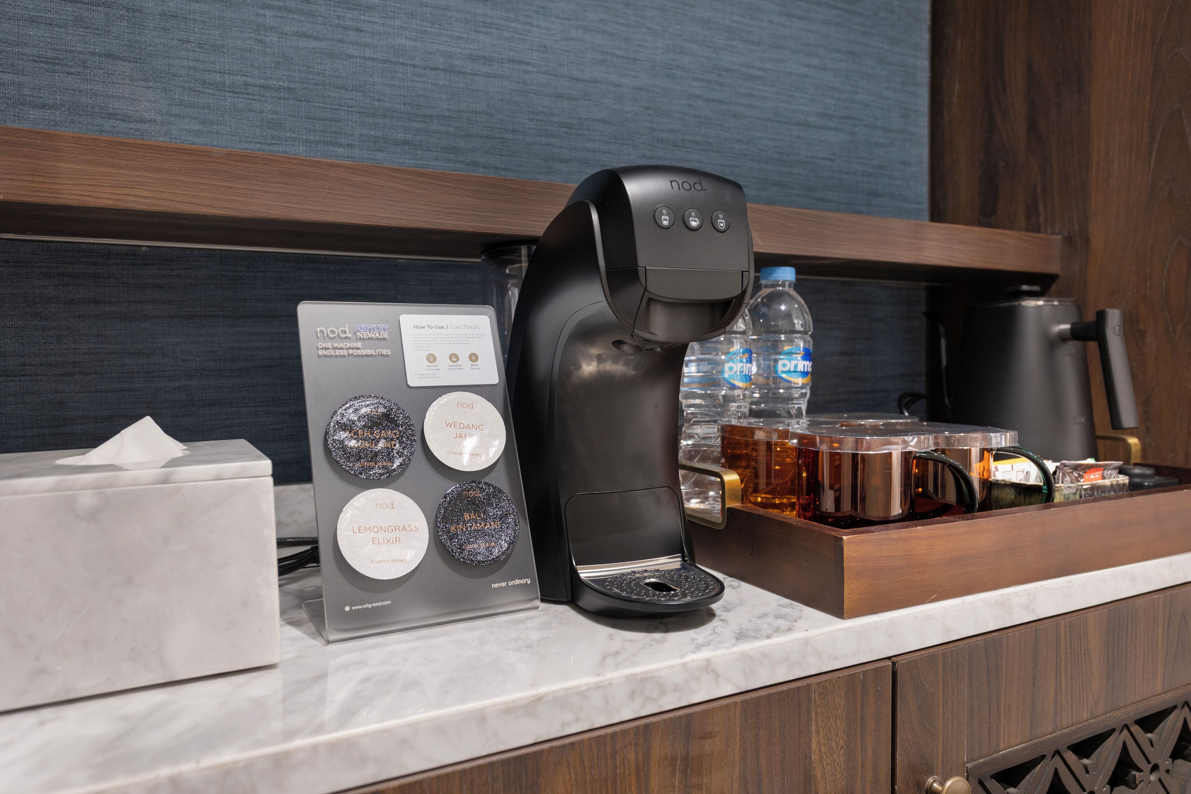 Uma cafeteira, uma garrafa de água, uma caixa de lenços e uma bandeja com diversos objetos em uma bancada de hotel.