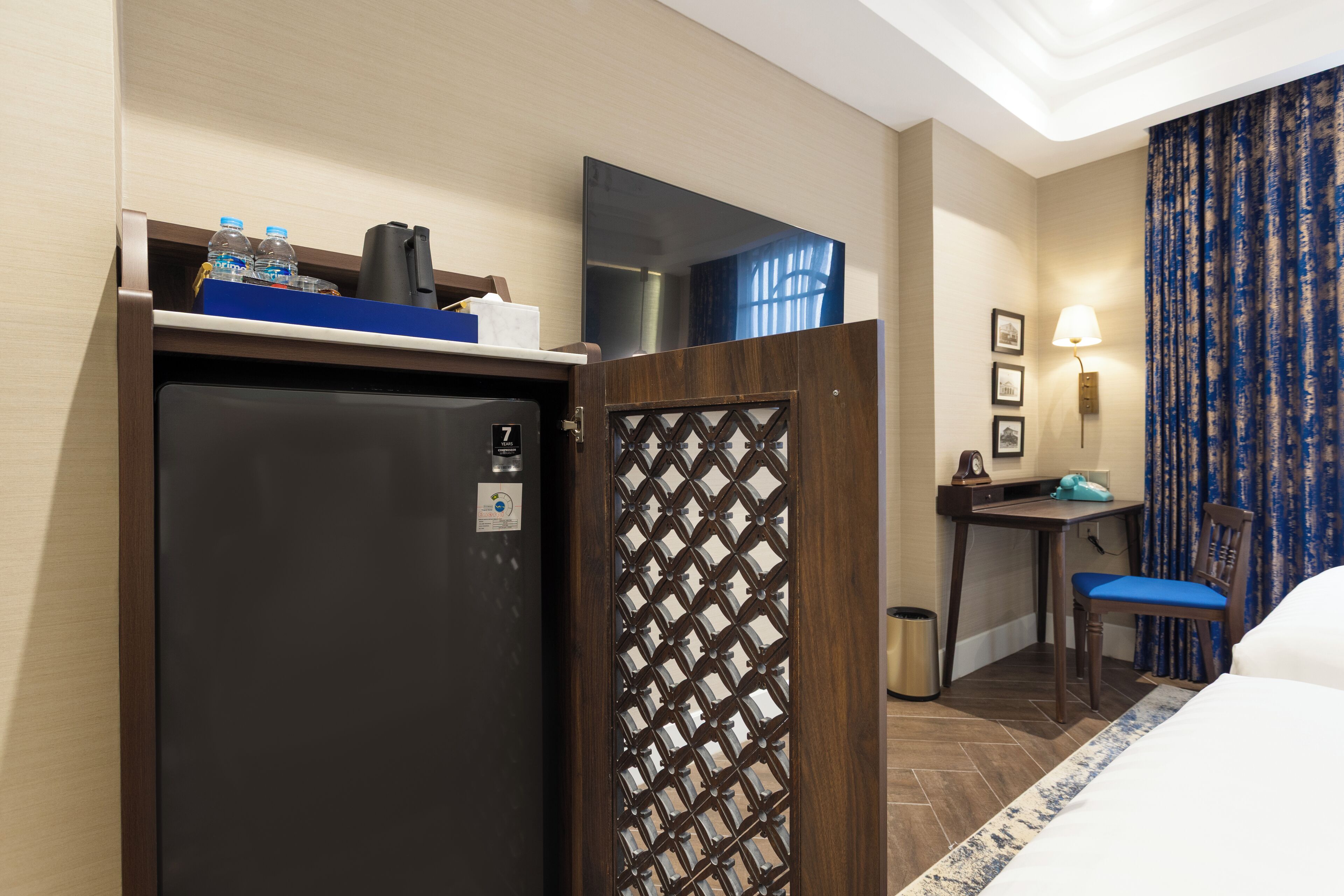 Une chambre d’hôtel équipée d’un lit, d’un bureau, d’une chaise, d’un réfrigérateur, d’une poubelle et d’une fenêtre avec des rideaux.