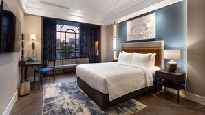 Habitación de hotel con una cama grande, un escritorio con una silla, una mesita con lámpara, un cesto de basura y un ventanal con cortinas.