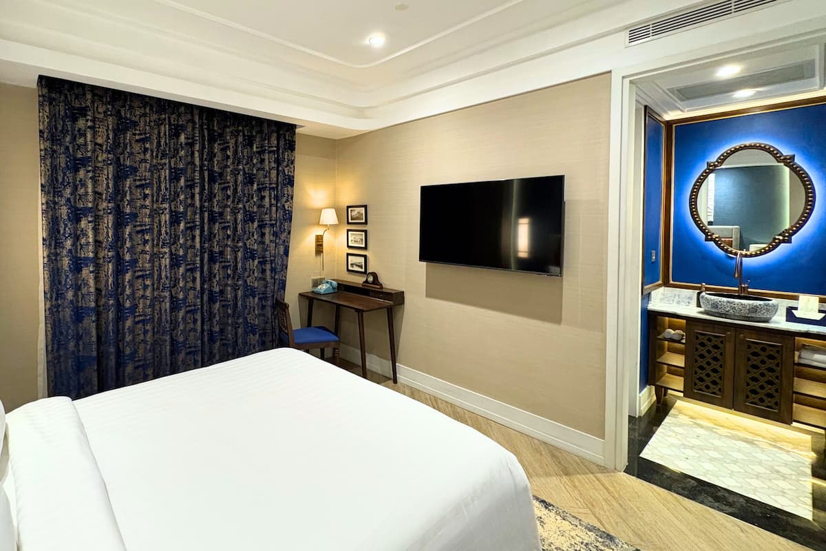 Habitación de hotel con una cama grande, un escritorio con silla, un televisor de pantalla plana, un baño con espejo y una lámpara en la pared.