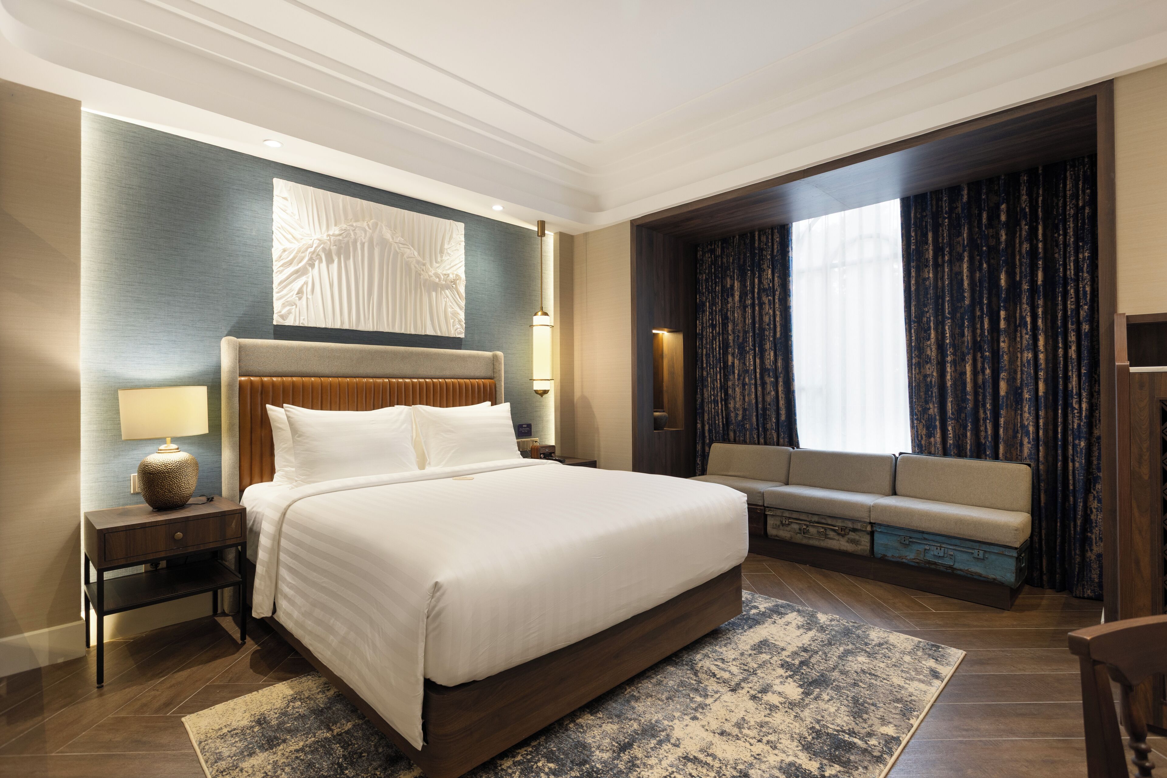 A modern hotel room with a large bed, a sofa, a nightstand, and a wall-mounted lamp.