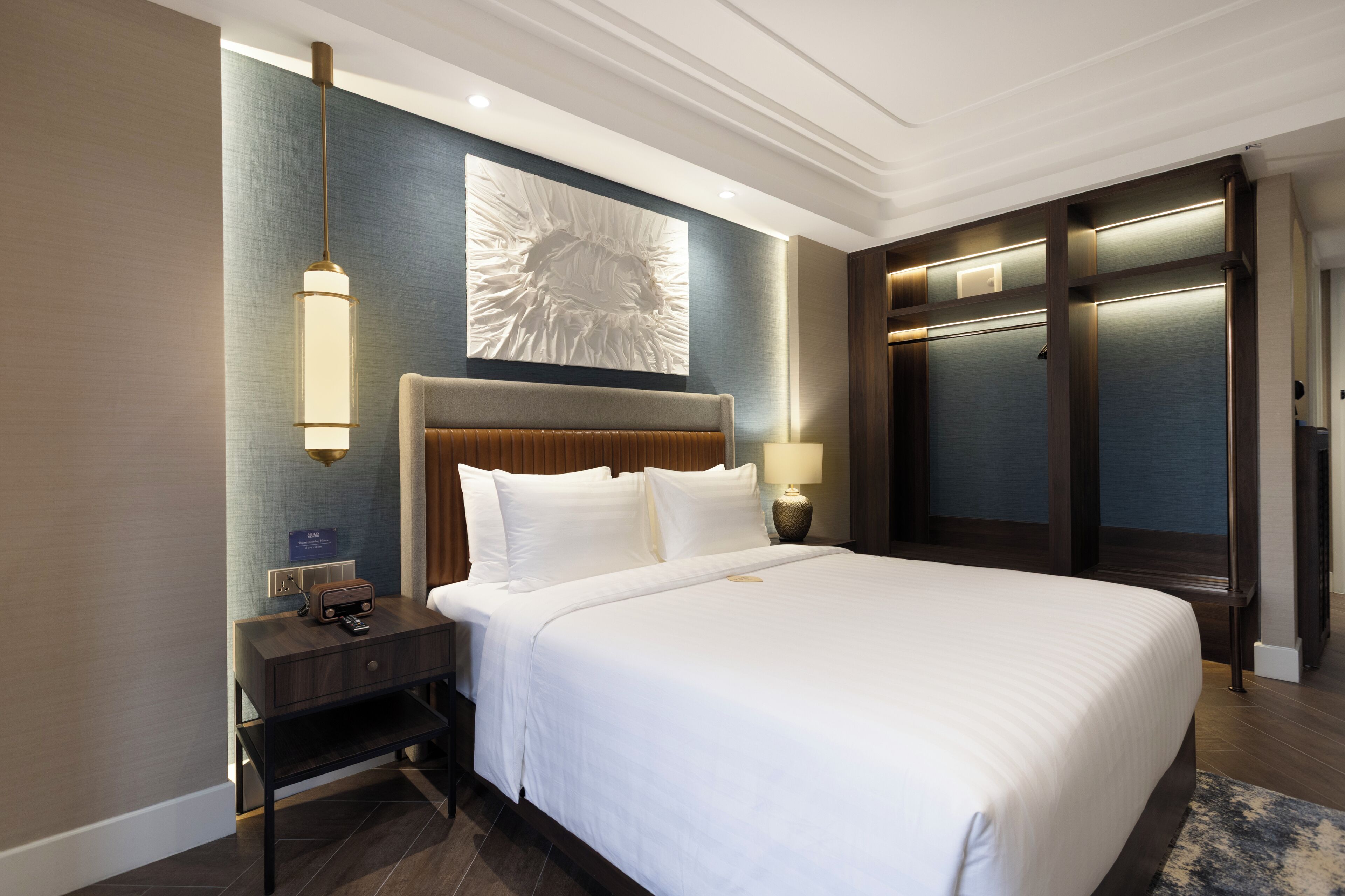 A hotel room with a large bed, a nightstand, a lamp, a wall art piece, and a wardrobe.