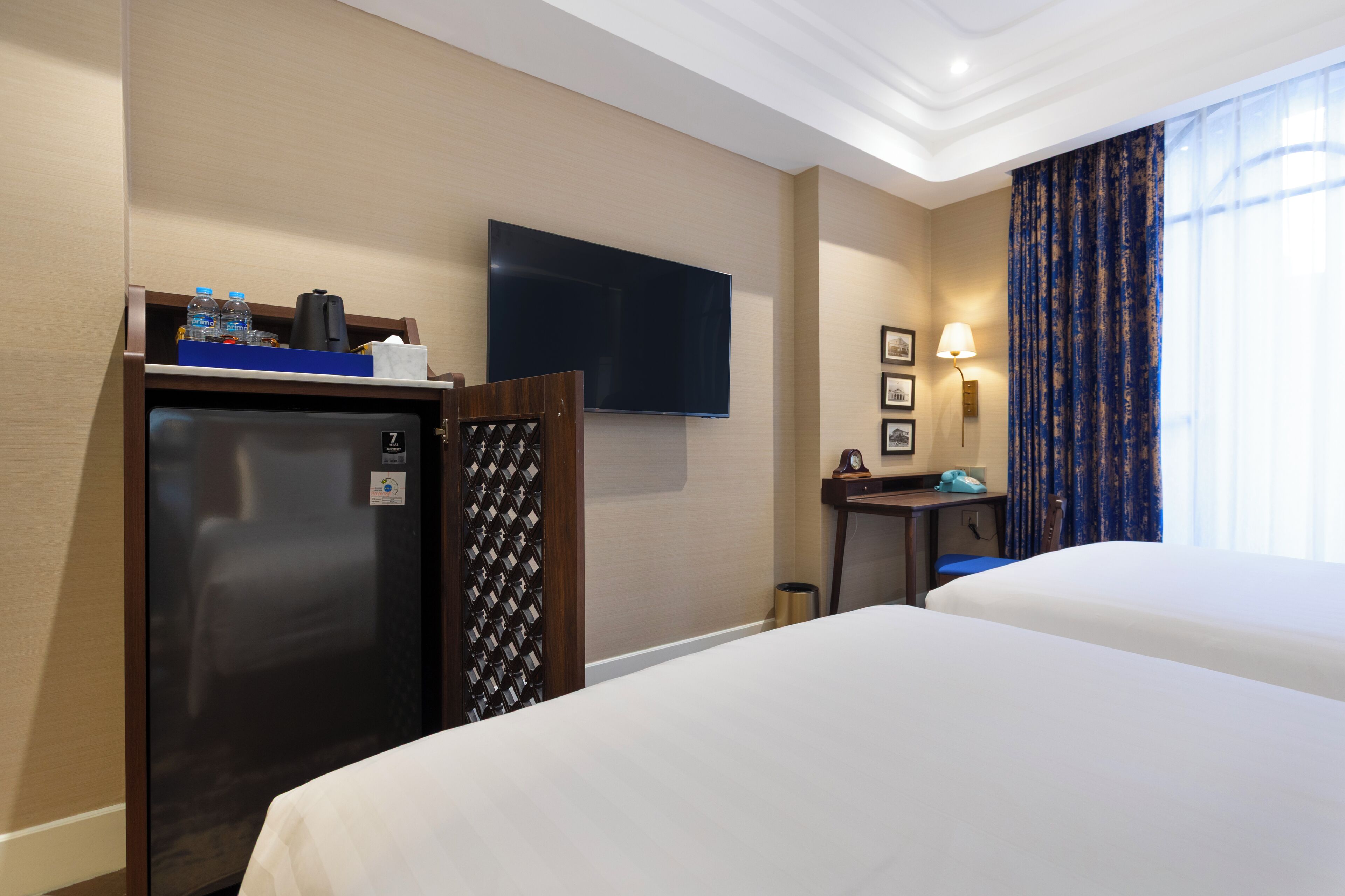 Une chambre d’hôtel avec deux lits, un téléviseur à écran plat, un minibar, un bureau avec une lampe et une fenêtre avec des rideaux.