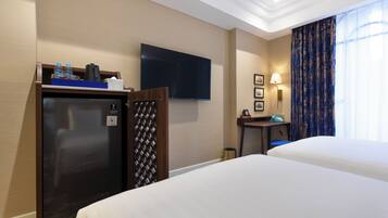 A hotel room with two beds, a flat-screen TV, a minibar, a desk with a lamp, and a window with curtains.
