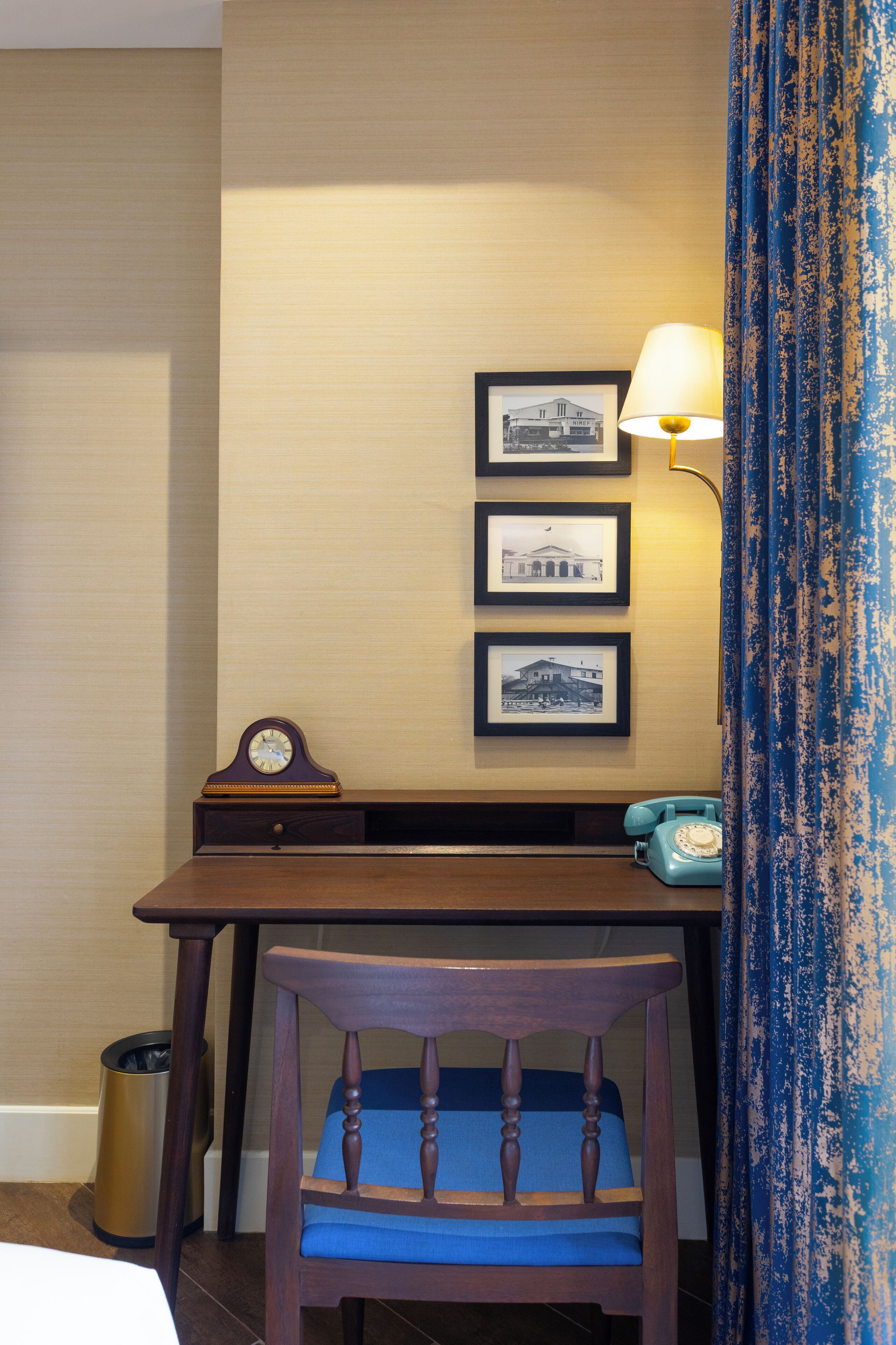A hotel room with a wooden desk, a blue chair, a lamp, and framed pictures on the wall.