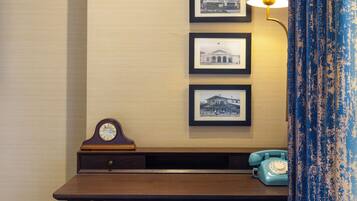 A hotel room with a wooden desk, a blue chair, a lamp, and framed pictures on the wall.