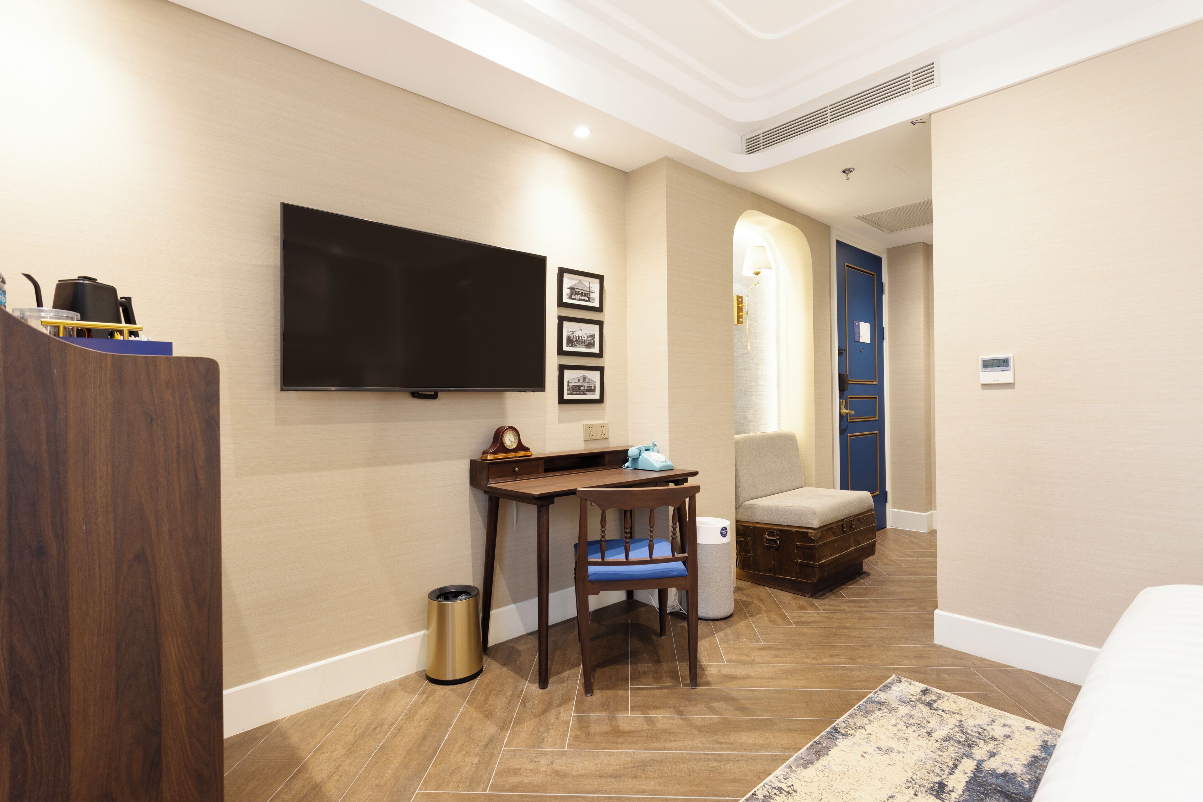 A hotel room with a flat-screen TV mounted on the wall, a desk with a chair, a trash can, and a small seating area with a cushioned bench.