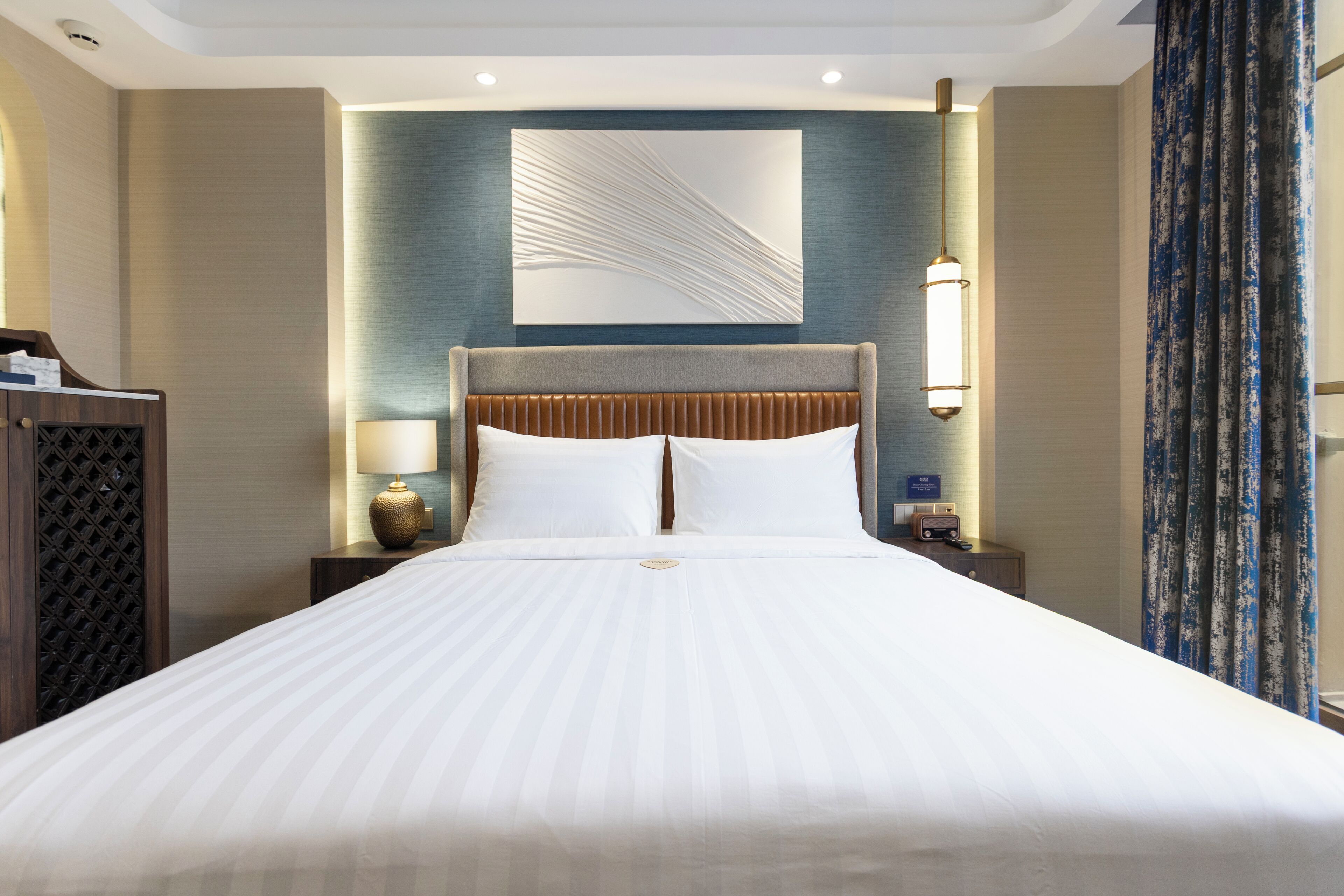 Een hotelkamer met een groot bed, een hoofdbord met verticale latten, een wanddecoratie, een bedlamp en een nachtkastje met een radio.