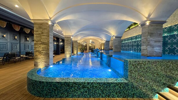 Una amplia piscina interior con diseño de mosaicos, rodeada de pilares de piedra y suelo de madera.