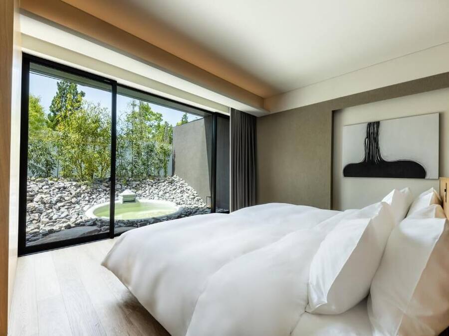 A modern bedroom with a large bed, a view of a garden through floor-to-ceiling windows, and a minimalist decor.