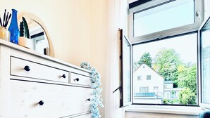 Une pièce avec une commode blanche, une fenêtre donnant sur des maisons et une chaise bleue.