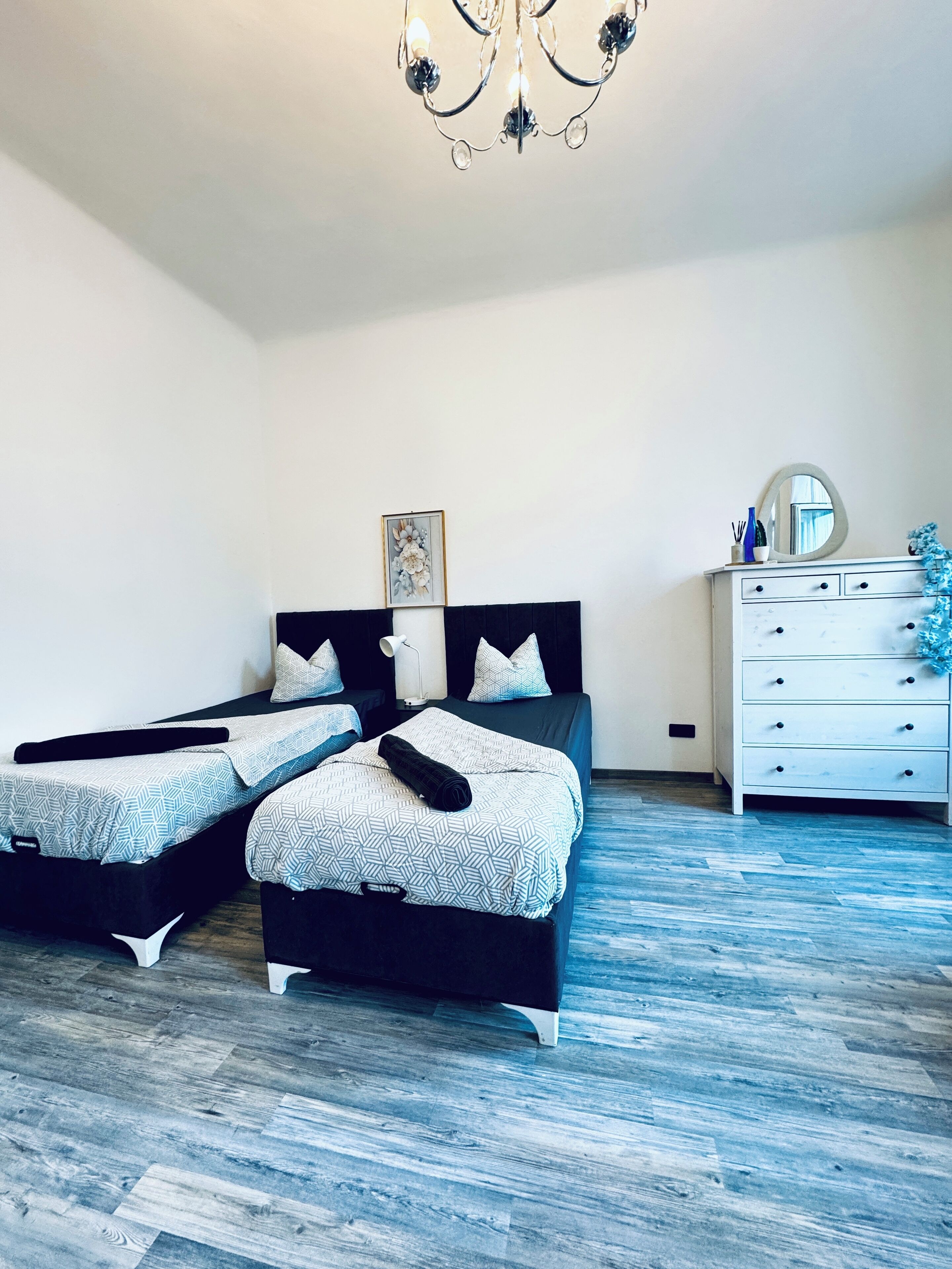 A bedroom with two beds, a dresser, a chandelier, and a framed picture on the wall.