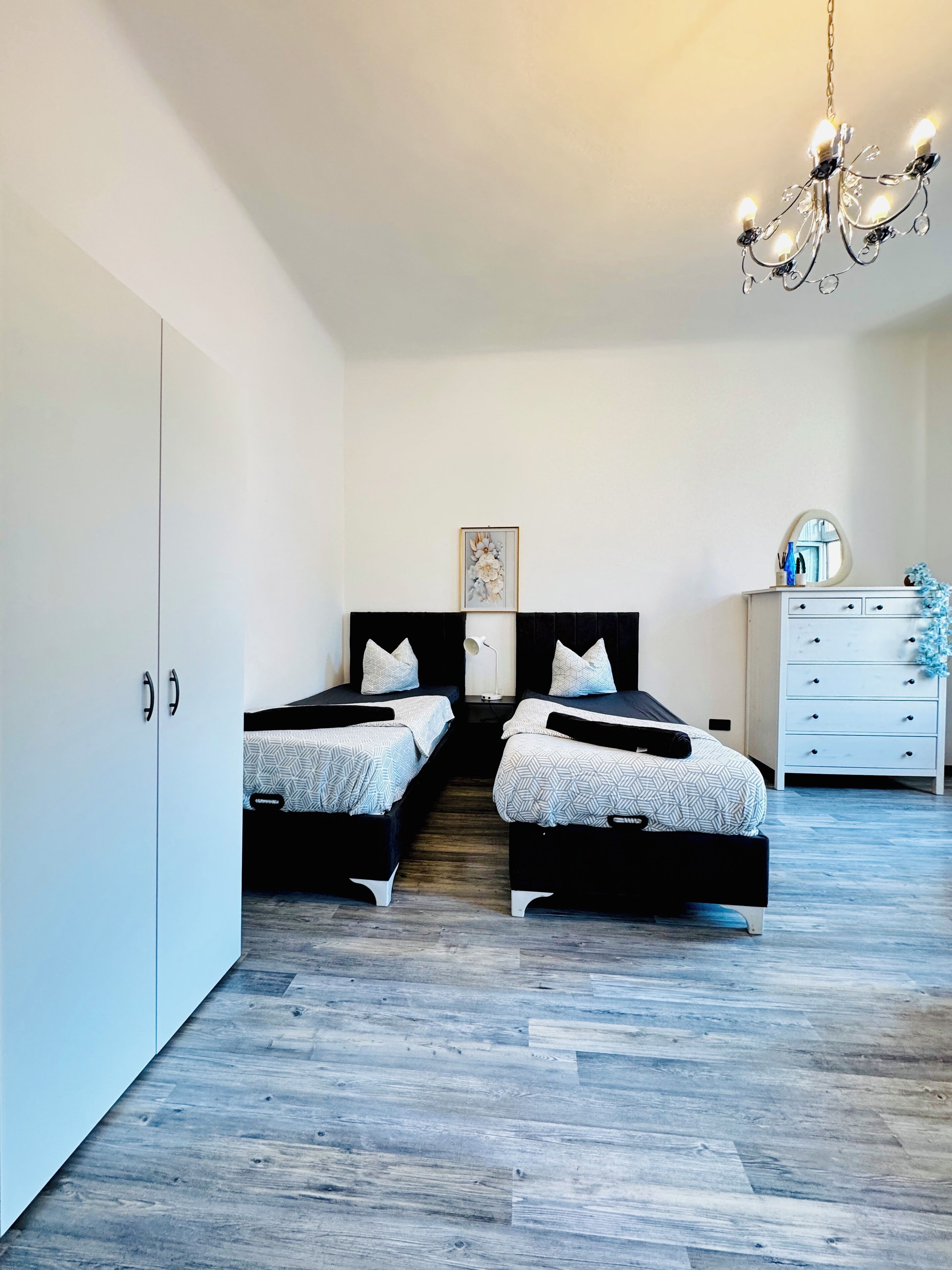 A bedroom with two beds, a dresser, a chandelier, and a framed picture on the wall.