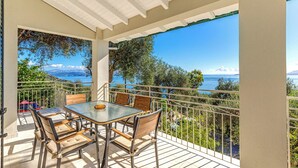Un balcon avec une table et des chaises en verre, offrant une vue panoramique sur la mer et les montagnes.