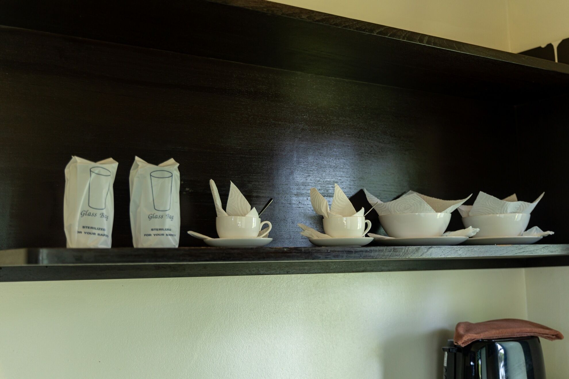 A kitchen shelf with two glass bags labeled 'Glass Bag' and a set of white ceramic cups and saucers.