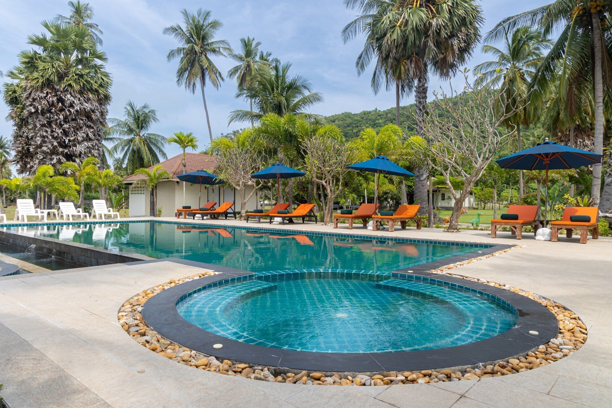 A pool area with lounge chairs and umbrellas, surrounded by palm trees and a mountainous backdrop.