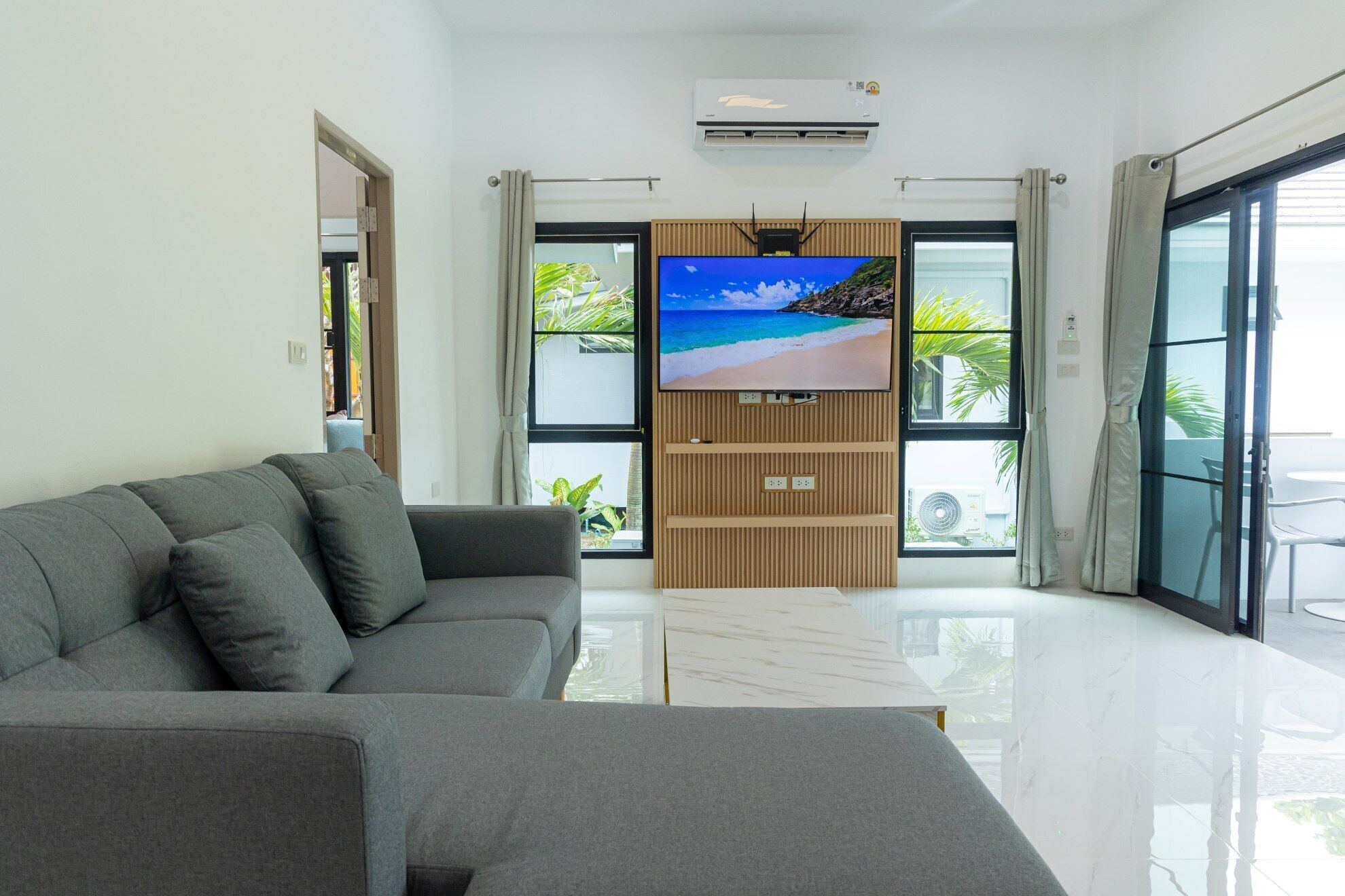 Ein modernes Wohnzimmer mit grauem Sofa, einem Flachbildfernseher an einer Holzvertäfelung und großen Fenstern mit Blick ins Freie.