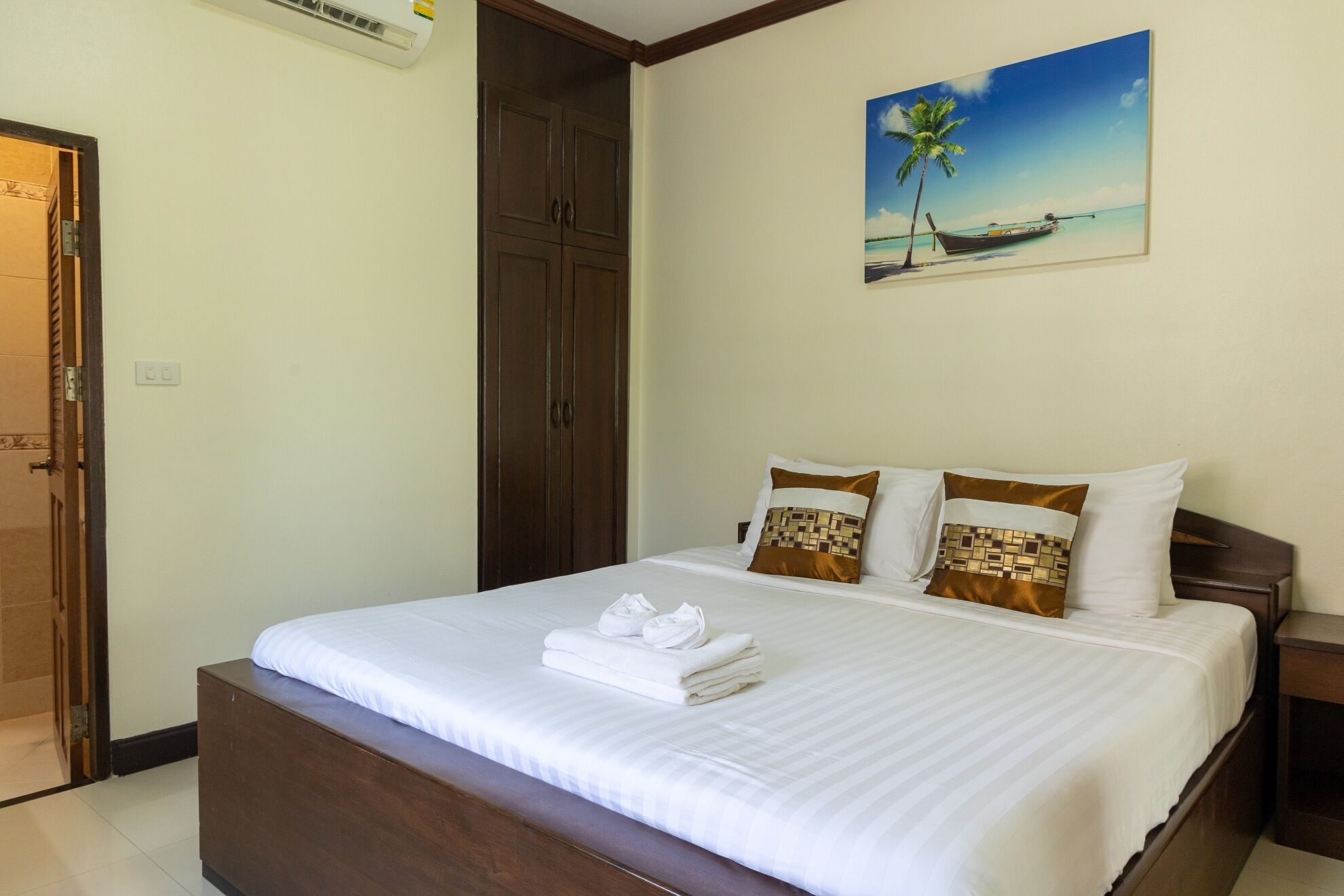 A neatly made bed with white linens and two decorative pillows, a wooden wardrobe, and a painting of a tropical beach scene on the wall.