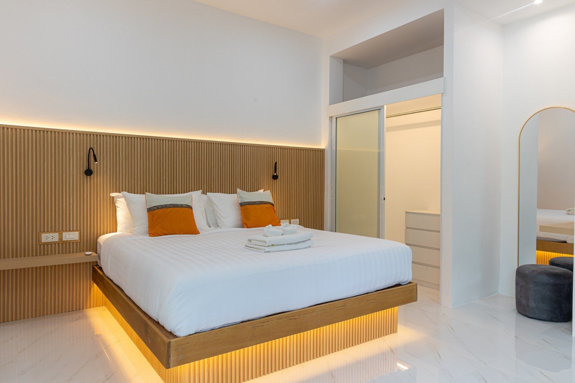 Ein modernes Hotelzimmer mit einem Holzkopfende, einem großen Bett mit weißer und orangefarbener Bettwäsche, einem Nachttisch mit Lampe und einem Kleiderschrank mit Spiegel.