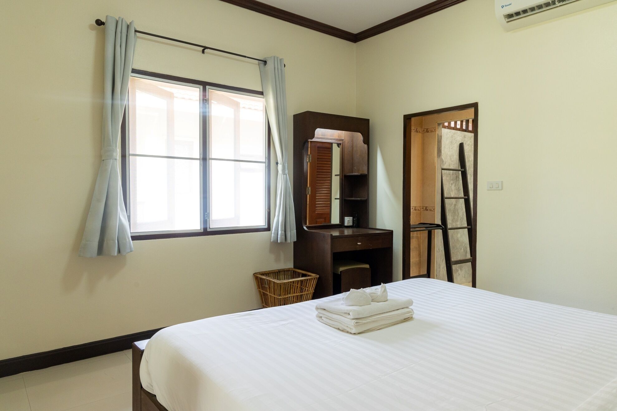 A hotel room with a bed, a wardrobe, a mirror, a window with curtains, a small wicker basket, and a ladder.