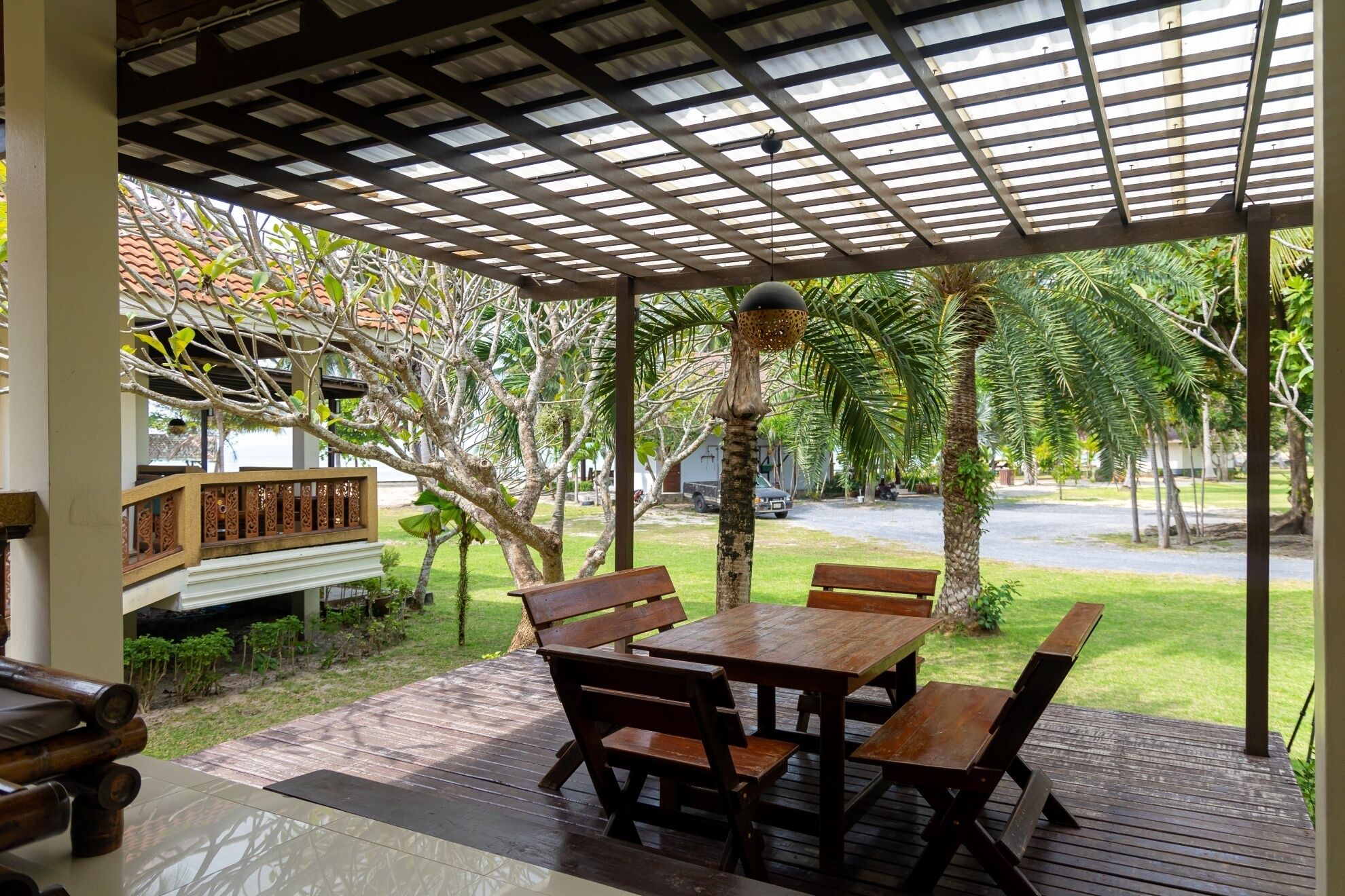 An outdoor patio with a wooden table and benches, surrounded by lush greenery and a covered area with a metal roof.