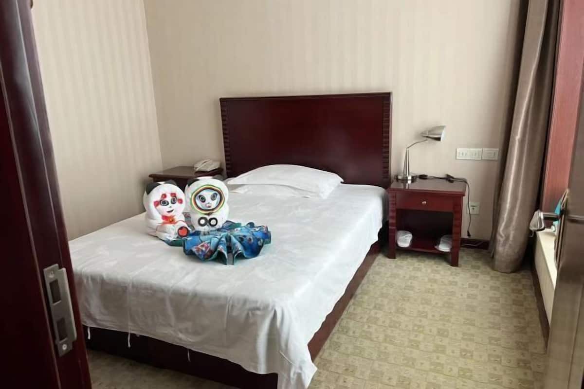 A hotel room with a single bed, a bedside table with a lamp, a small shelf with shoes, and a wall socket.