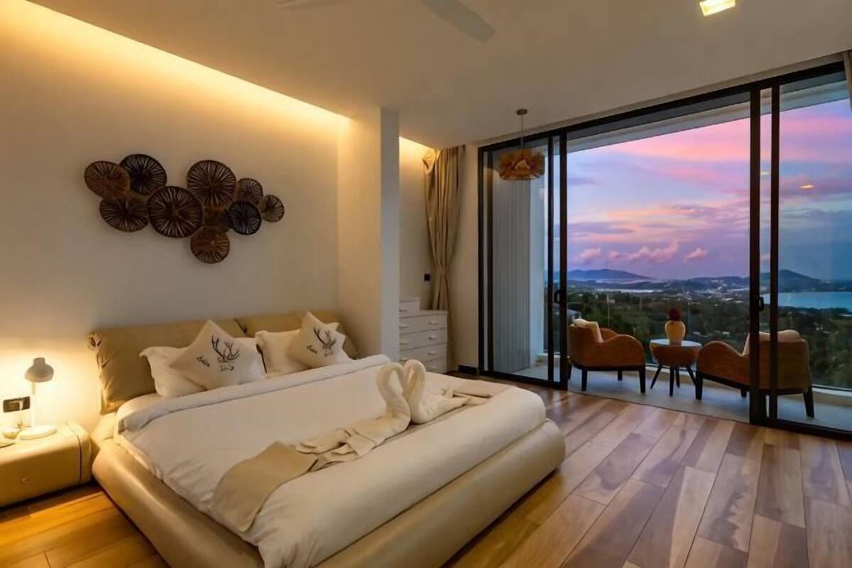 A bedroom with a large bed, a view of the cityscape, and a balcony with chairs.