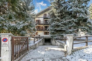 Un sendero cubierto de nieve que conduce a un edificio de varios pisos con balcones, rodeado de árboles cargados de nieve.