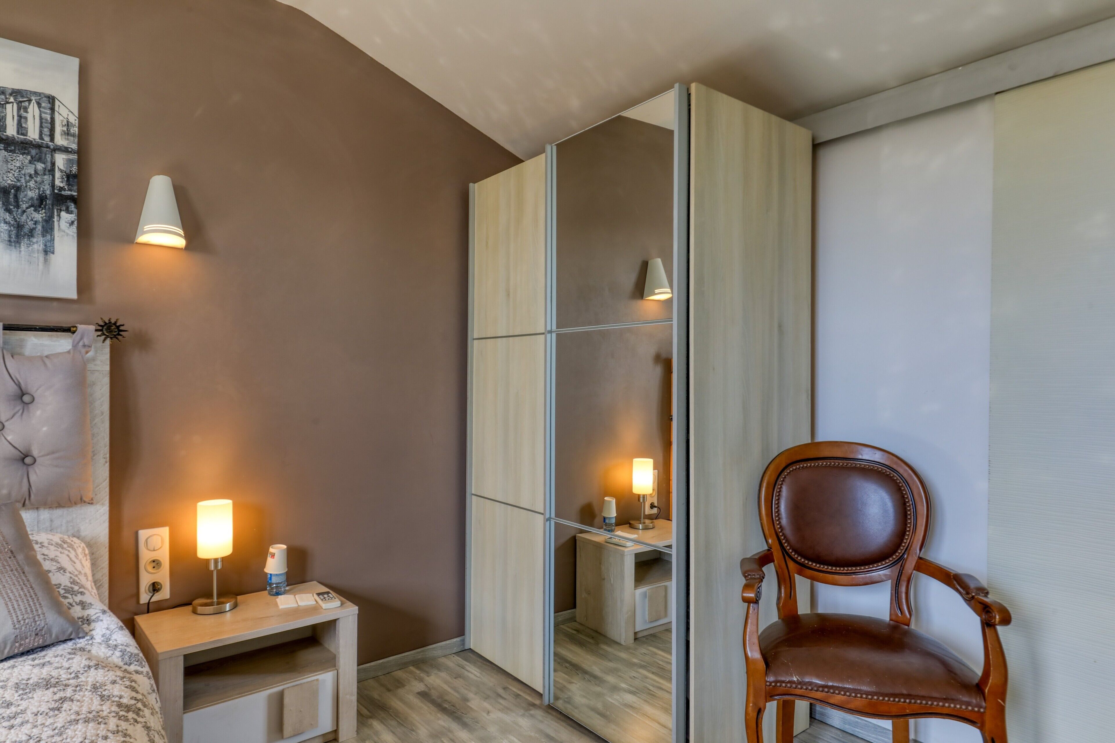 A bedroom with a bed, bedside table, lamp, chair, and a large mirror.