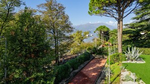 A pathway bordered by lush greenery, a large tree, and a distant mountain view. - Lakefront Serenity Villa With Pool (Mandello del Lario)