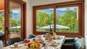 A dining table set with breakfast items, including fruits, pastries, and coffee, in a room with large windows overlooking a mountainous landscape. - Lakefront Serenity Villa With Pool (Mandello del Lario)