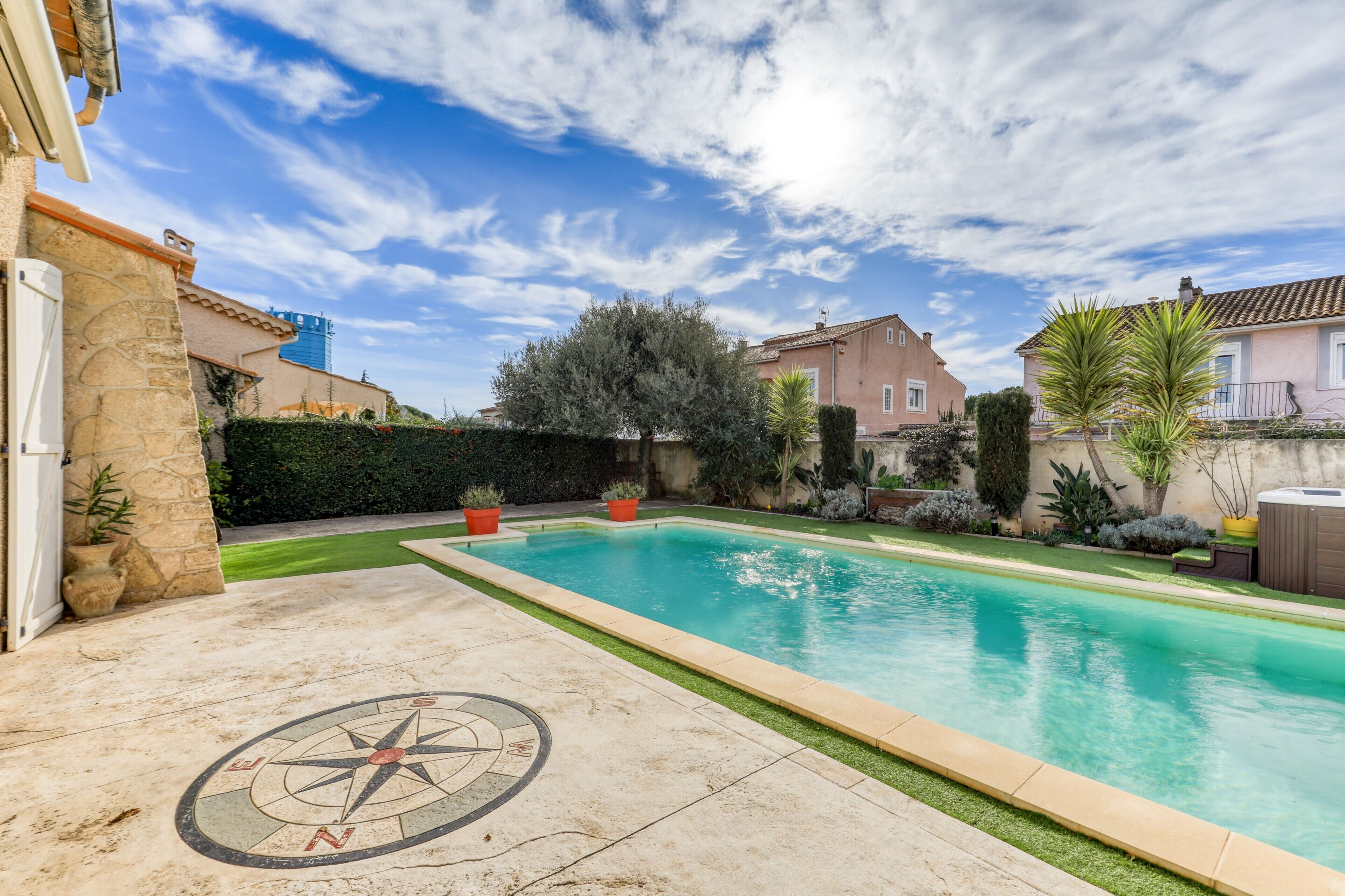 A swimming pool with a compass design on the ground, surrounded by greenery and houses.