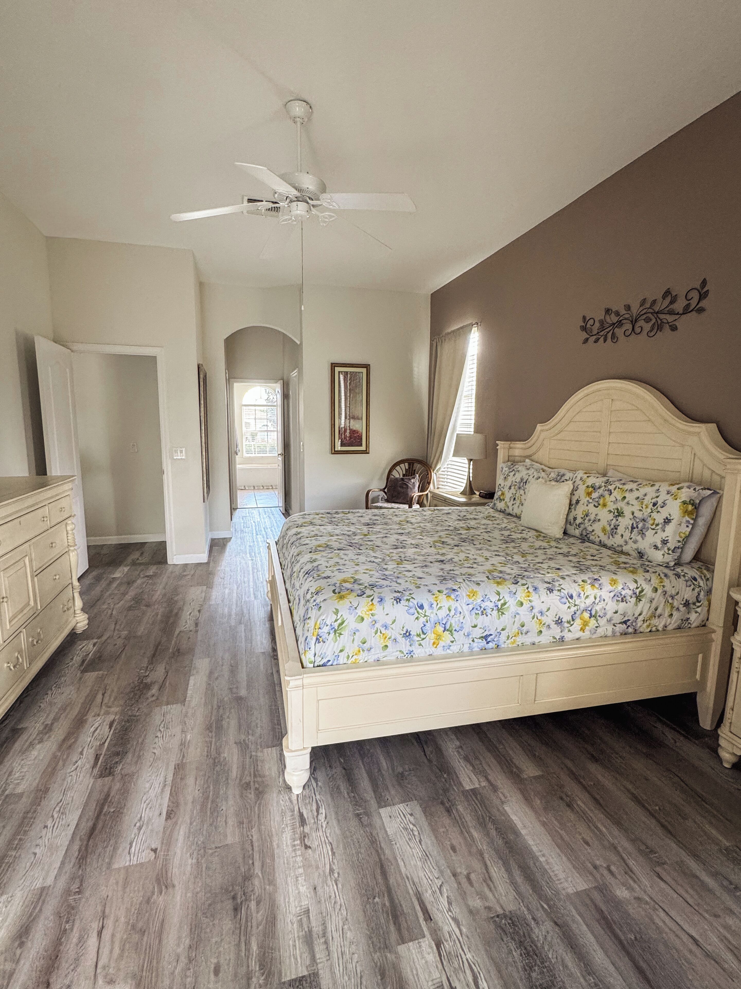 A bedroom with a large bed, a ceiling fan, a dresser, and a door leading to another room.