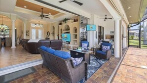 A spacious living area with a large sofa, a coffee table, and a television. There are ceiling fans and a view of the outdoors.
