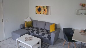 A modern living room with a grey sofa, a white coffee table, and a grey armchair. A sunflower painting adorns the wall, and a small shelf with a vase and calendar is visible. - Holiday Home 'Stahlberg' with Private Terrace, Balcony and Wi-Fi (Potsdam)