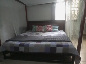 Una cama con colcha y almohadas, una ventana con cortinas, y vista al exterior.